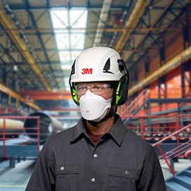 Hombre con máscara de seguridad, gafas protectoras, casco y orejeras. Sala de fábrica al fondo.